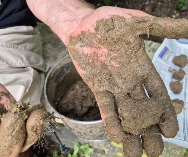 Eine Hand hält eine erdige, unregelmäßig geformte Kugel. Die Finger und die Handfläche sind mit feuchter Erde bedeckt. Im Hintergrund stehen ein Metallgefäß mit Erde und eine Zeitung auf einer Holzfläche. Auf der Zeitung liegen mehrere weitere erdige Kugeln.