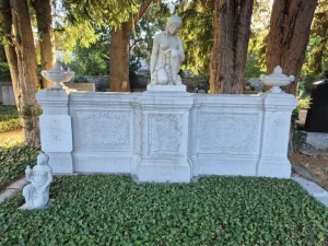 Ein weißes Grabmal aus Stein steht vor mehreren hohen Bäumen. Das Denkmal besteht aus einer rechteckigen Wand mit floralen Reliefs und Inschriften. Auf dem Mittelteil kniet eine größere Skulptur einer nachdenklichen, halb entkleideten Person. Links auf dem Boden befindet sich eine kleinere Skulptur einer knienden Figur mit gefalteten Händen. Die Fläche vor dem Grabmal ist mit dichtem, grünem Bodendecker bewachsen. Im Hintergrund sind weitere Grabsteine und Bäume zu sehen.