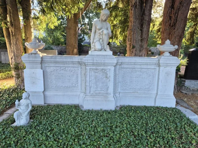 Ein weißes Grabmal aus Stein steht vor mehreren hohen Bäumen. Das Denkmal besteht aus einer rechteckigen Wand mit floralen Reliefs und Inschriften. Auf dem Mittelteil kniet eine größere Skulptur einer nachdenklichen, halb entkleideten Person. Links auf dem Boden befindet sich eine kleinere Skulptur einer knienden Figur mit gefalteten Händen. Die Fläche vor dem Grabmal ist mit dichtem, grünem Bodendecker bewachsen. Im Hintergrund sind weitere Grabsteine und Bäume zu sehen.