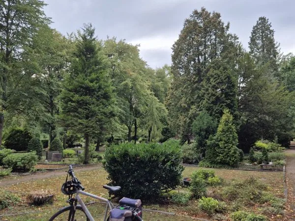 Im Vordergrund steht ein Fahrrad mit Gepäcktasche auf einem Weg. Dahinter erstreckt sich eine gepflegte Grünanlage mit verschiedenen Büschen, Bäumen und Grabsteinen. Die Szene wirkt ruhig und ist von dichtem, grünem Bewuchs geprägt. Der Himmel ist bewölkt.