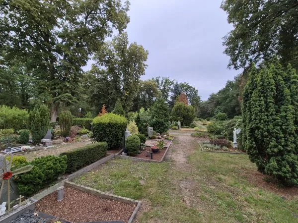 Ein schmaler Weg führt durch eine gepflegte Friedhofsanlage mit Grabsteinen, niedrigen Hecken und verschiedenen immergrünen Sträuchern. Einzelne Gräber sind mit Blumen und kleinen Dekorationen geschmückt. Im Hintergrund stehen hohe Laubbäume, der Himmel ist bewölkt.