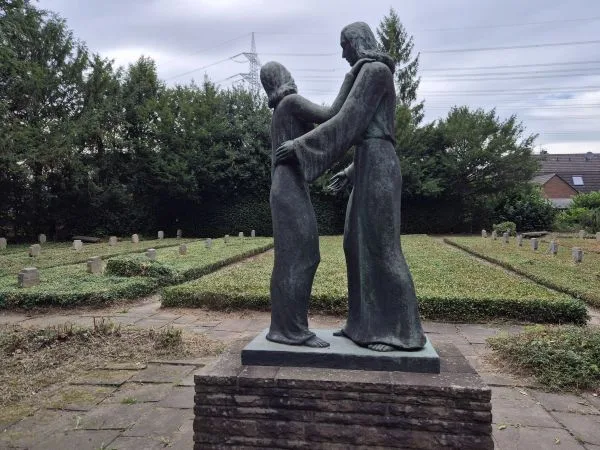 Im Vordergrund steht eine bronzefarbene Skulptur auf einem niedrigen Sockel aus Backstein. Die Skulptur zeigt zwei menschliche Figuren in langer Gewandung, die sich gegenüberstehen und einander an den Schultern halten. Im Hintergrund sind gepflegte Grabreihen mit niedrigen Grabsteinen zu sehen, die von Hecken und Bäumen umgeben sind. Der Himmel ist bewölkt, und im Hintergrund verlaufen Stromleitungen über das Bild.