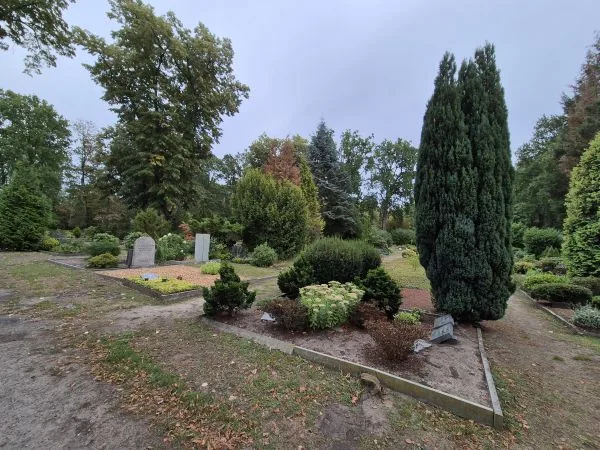 Ein gepflegter Friedhof mit mehreren Grabsteinen und dicht bepflanzten Gräbern. Verschiedene immergrüne und laubabwerfende Bäume sowie Sträucher umrahmen die Anlage. Die Wege sind mit hellem Kies bedeckt, und einzelne Beete sind von niedrigen Hecken eingefasst. Der Himmel ist bewölkt.