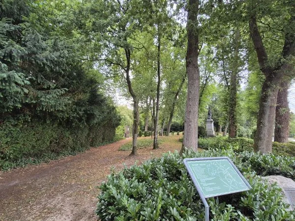 Im Vordergrund steht eine grüne Informationstafel mit weißer Schrift und Linienzeichnung, die von dichten, grünen Sträuchern umgeben ist. Dahinter verläuft ein unbefestigter Weg durch einen Friedhof, der von hohen Bäumen und Hecken gesäumt wird. Im Hintergrund sind Grabsteine und Denkmäler zwischen den Bäumen zu erkennen. Das Licht fällt durch das dichte Blätterdach und erzeugt eine ruhige Atmosphäre.