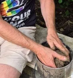 Zwei Hände formen eine erdige, grobkörnige Mischung in einem Metaltopf. Die Person trägt ein schwarzes T-Shirt mit buntem Aufdruck und eine helle kurze Hose und sitzt auf einer Steinbank. ImHintergrund sind grüne Blätter zu sehen.