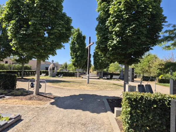 Eingang zum Freidhof Morschenich mit mehreren Bäumen, die Schatten auf die Wege werfen. In der Mitte steht ein großes Holzkreuz. Im Hintergrund sind moderne Steinbänke und Skulpturen zu sehen. Die Wege sind mit Kies bedeckt und von niedrigen Hecken eingefasst. Der Himmel ist wolkenlos und blau.