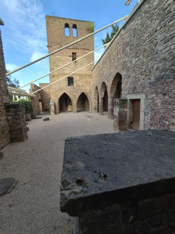 Im Vordergrund steht ein rechteckiger Altar aus Stein, dahinter erstreckt sich ein Innenhof mit Kiesboden. Die Umfassungsmauern bestehen aus grobem Naturstein und zeigen mehrere Rundbogenöffnungen. Links erhebt sich ein hoher, rechteckiger Turm mit zwei schmalen Fensteröffnungen. Über den Hof verlaufen helle Metallstreben, die sich zwischen den Mauern und dem Turm spannen. Der Himmel ist blau mit wenigen Wolken.
