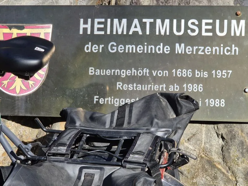 Das Bild zeigt ein Schild des Heimatmuseums der Gemeinde Merzenich. Auf dem Schild steht: HEIMATMUSEUM der Gemeinde Merzenich Bauerngehöft von 1686 bis 1957 Restauriert ab 1986 Fertiggestellt 1988 Davor ist ein schwarzes Fahrrad abgestellt.