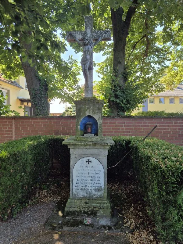 Im Vordergrund steht ein steinernes Grabmal mit einem Kreuz und einer Christusfigur. Das Grabmal ist von einer niedrigen Hecke eingefasst und befindet sich vor einer roten Backsteinmauer. Im Hintergrund sind gelbe Gebäude und hohe Bäume zu sehen, deren Blätter Schatten auf das Grabmal werfen. Auf dem Sockel des Grabmals ist eine Inschrift eingraviert, die teilweise lesbar ist. In einer kleinen Nische unterhalb des Kreuzes steht eine Kerze.