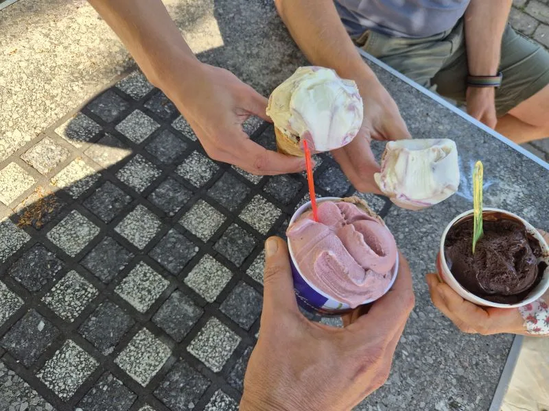 Auf dem Bild sieht man mehrere Personen im Freien, die Eisbecher und Eiswaffeln halten. Oben links und oben rechts halten zwei Personen jeweils eine Eiswaffel mit hellem Eis. Unten in der Mitte hält eine Person einen Becher mit rosa Eis und rotem Löffel. Unten rechts hält eine Person einen Becher mit dunklem Schokoladeneis und gelbem Löffel. Die Gruppe sitzt an einem Steintisch mit Schachbrettmuster im Freien.