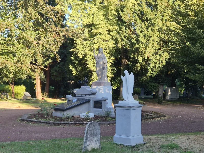 Im Vordergrund steht eine helle Engelsstatue auf einem Sockel. Dahinter befindet sich ein großes Grabmal mit einer stehenden weiblichen Figur, die einen langen Mantel trägt. Die Szene ist von zahlreichen Bäumen umgeben, deren dichtes Laub Schatten auf den Boden wirft. Der Friedhof wirkt gepflegt, mit klar abgegrenzten Wegen und einzelnen Grabsteinen.