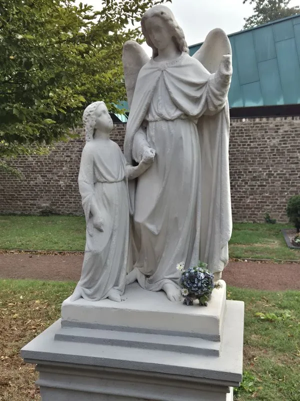 Im Vordergrund steht eine helle, steinerne Statue auf einem Sockel. Die Figur zeigt einen Engel mit ausgebreiteten Flügeln, der eine kleinere, kindliche Gestalt an der Hand hält. Beide Figuren tragen lange Gewänder. Am Sockel steht eine Steintroll-Figur mit großen Augen. Aus den Steinporen wachen Blumen, Gras und Efeu.. Im Hintergrund sind eine niedrige Mauer aus hellen Ziegeln, ein grüner Rasen und Bäume zu sehen.