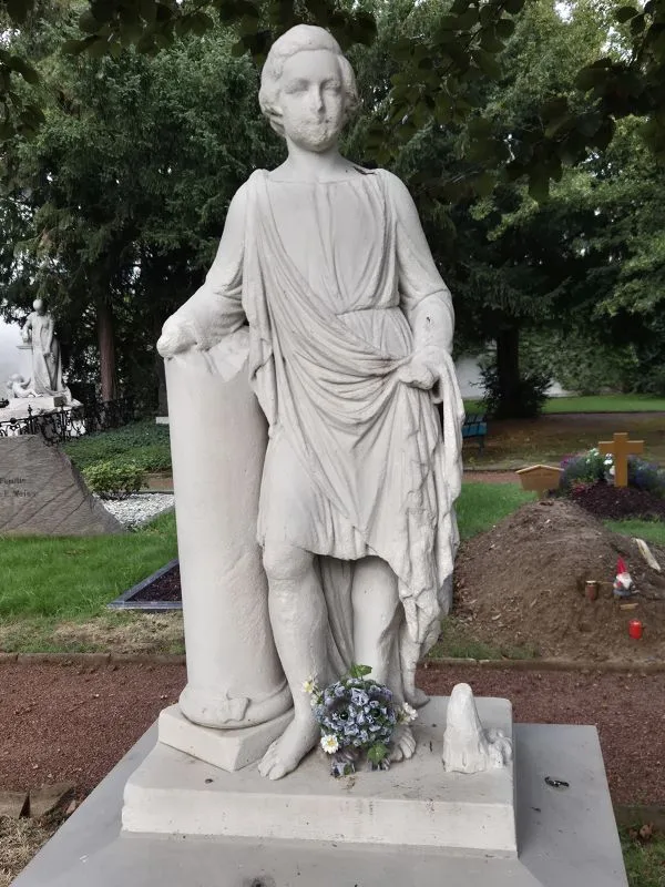 Eine lebensgroße Statue aus hellem Stein steht aufrecht auf einem Sockel. Die Figur trägt ein antik anmutendes, locker drapiertes Gewand und lehnt mit dem rechten Arm an einer Säule. Am Sockel steht eine Steintroll-Figur mit großen Augen. Aus den Steinporen wachen Blumen, Gras und Efeu. Im Hintergrund sind Grabkreuze, Grabsteine und gepflegte Grünflächen zu sehen. Bäume spenden Schatten über die Szene.