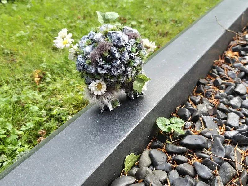 Auf einer steinernen Umrandung steht eine kleine, künstliche Figur, die mit grauen Steinen und Moos bedeckt ist und große, grüne Augen hat. Die Figur ist von künstlichen Blättern, weißen Blüten und Zweigen umgeben. Die Figur blickt auf eine mit Steinschotter gefüllte Fläche. Im Hintergrund ist eine Wiese erkennbar.