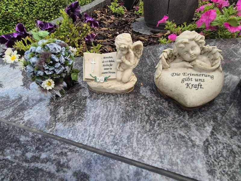 Auf einer steinernen Platte steht eine kleine, künstliche Figur, die mit grauen Steinen und Moos bedeckt ist und große, grüne Augen hat. Die Figur ist von künstlichen Blättern, weißen Blüten und Zweigen umgeben. Daneben sind steinerne Engelfiguren.