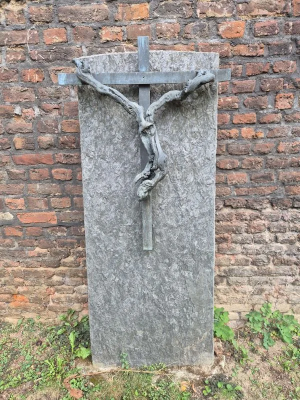 Ein schlichtes Kreuz mit einer knorrigen Reliefverzierung auf einer hellen Steinplatte steht vor vor einer roten Backsteinmauer.