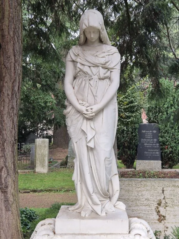 Eine weiße Statue einer trauernden Frau mit langen Gewändern steht inmitten von Bäumen auf einem Friedhof.