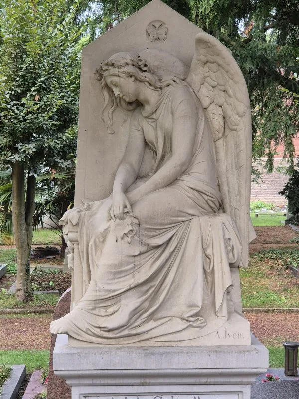Eine sitzende Engelsfigur aus Stein auf einem Grabmal in einer parkähnlichen Friedhofsanlage.