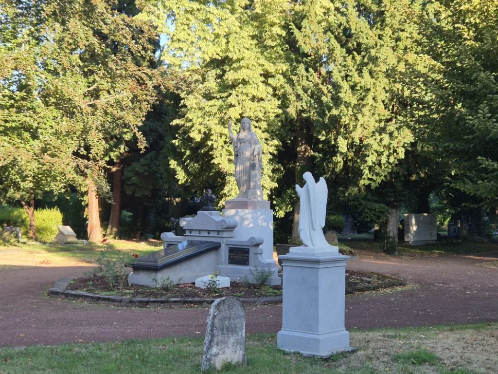 Mehrere Grabdenkmäler mit Figuren und eine Engelsskulptur stehen in einem sonnigen, baumbestandenen Friedhof.