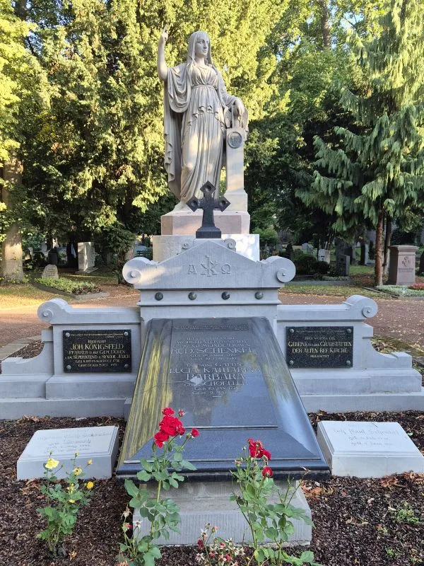 Ein aufwendig gestaltetes Grabmal mit einer weiblichen Statue und Blumen vor einer baumbestandenen Friedhofsanlage.