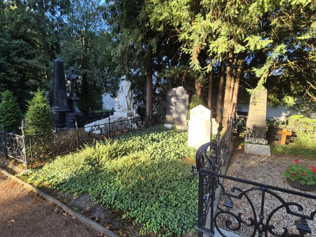 Mehrere Grabsteine und ein weißes Denkmal in einem eingezäunten, sonnigen Grabfeld unter hohen Bäumen.