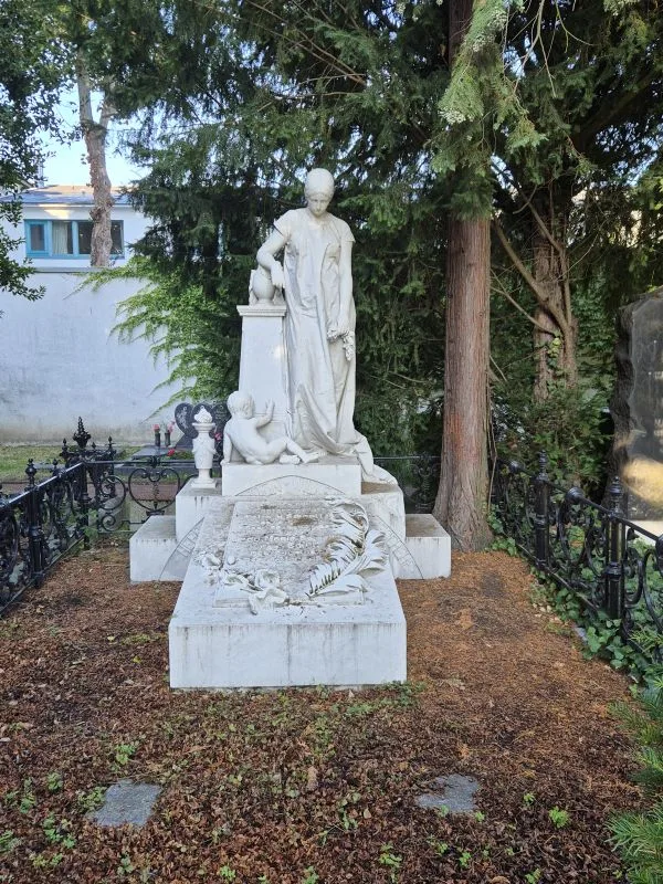 Ein weißes Grabmal mit einer stehenden Figur und einem kindlichen Figur in einem umfriedeten Grab.
