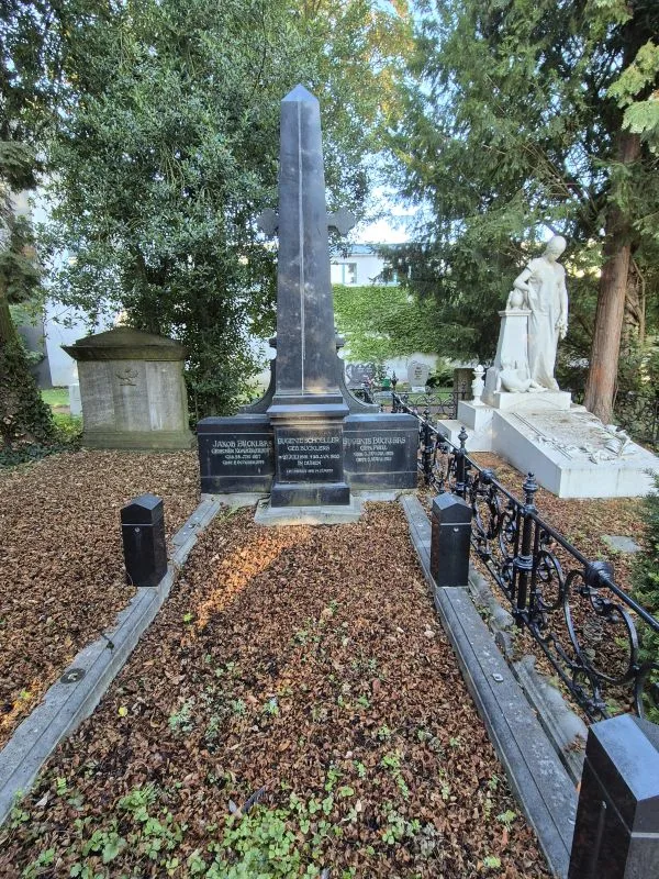 Ein hohes schwarzes Obelisken-Grabmal in einem von Zäunen umgebenen Grabfeld neben einer weißen Statue.