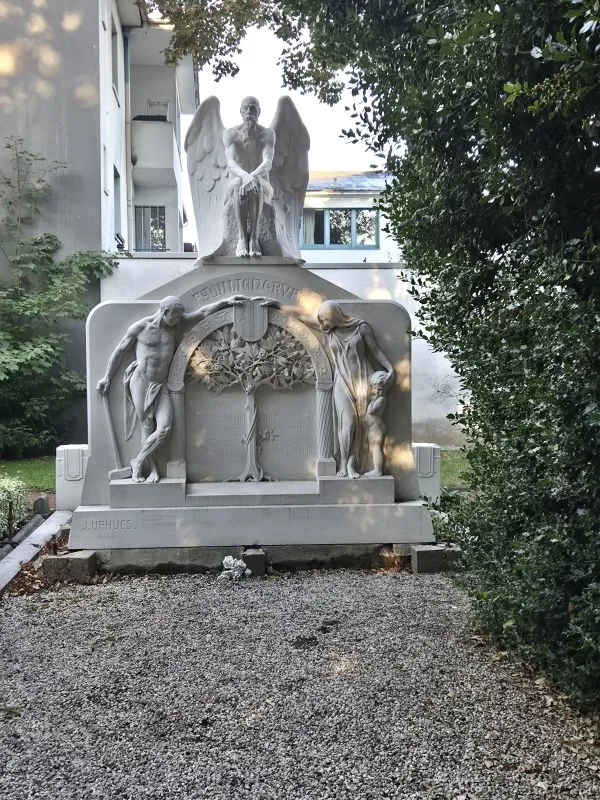 Ein großes, helles Grabmal aus Stein steht auf einem Kiesbett und wird von Bäumen und Sträuchern umrahmt. Das Denkmal zeigt mittig eine geflügelte Engelsfigur mit ausgebreiteten Armen, die auf einem Sockel steht. Links und rechts davon sind zwei stehende, unbekleidete männliche Figuren in nachdenklicher Haltung dargestellt. In der Mitte des Reliefs befindet sich ein stilisierter Lebensbaum. Oberhalb des Reliefs ist ein Schriftzug in den Stein gemeißelt. Im unteren Bereich ist eine weitere Inschrift zu erkennen.