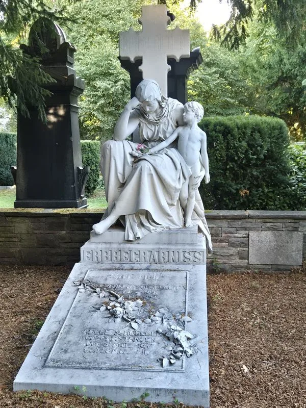 Im Vordergrund steht eine steinerne Grabskulptur, die eine sitzende Frau mit gesenktem Kopf und einen stehenden Jungen zeigt. Die Frau hält einen Blumenstrauß in der Hand, während der Junge sie sanft berührt. Die Skulptur befindet sich auf einem rechteckigen Grabstein, der mit Blumen und Inschriften versehen ist. Im Hintergrund sind weitere Grabmale und dichte grüne Bepflanzung zu sehen.