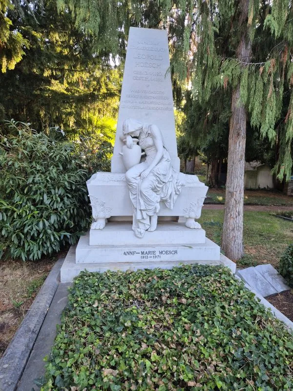 Im Vordergrund steht ein aufwendig gestaltetes, weißes Grabmal aus Stein. Eine sitzende Frauenfigur mit gesenktem Kopf und angewinkeltem Arm lehnt an einer Urne. Die Figur wirkt trauernd und ist in ein langes Gewand gehüllt. Das Grabmal trägt mehrere Inschriften, darunter einen Namen und Lebensdaten. Im unteren Bereich wächst dichter Efeu. Im Hintergrund stehen hohe Bäume und Sträucher, die das Grabmal teilweise beschatten.