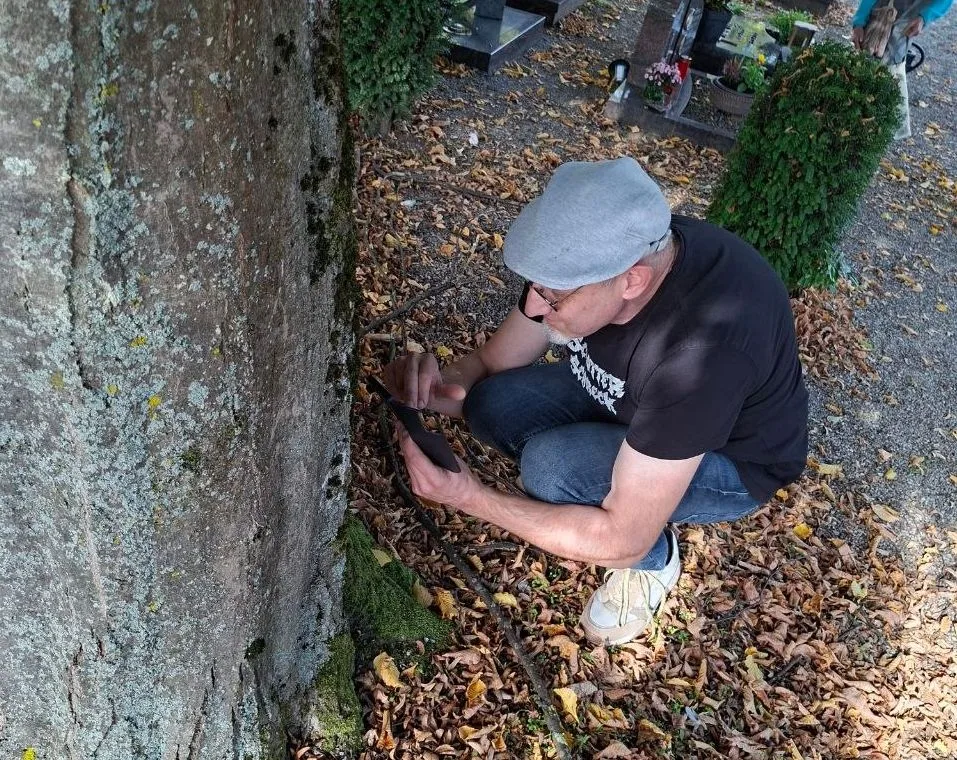 Ein Mann mit grauer Schirmmütze und schwarzem T-Shirt mit der Aufschrift Schotterschreck hockt neben einem Baumstamm auf einem Friedhof. Er hält ein Telefon in der Hand und richtet es auf den Baum. Der Boden ist mit trockenen Blättern bedeckt, im Hintergrund sind Grabsteine und Buchsbaumsträucher zu sehen.