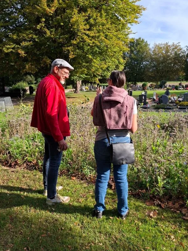 Zwei Personen stehen auf einer Wiese am Rand eines Friedhofs. Im Hintergrund sind Grabsteine und Gräber zu sehen. Die Person links trägt eine rote Jacke, eine helle Hose und eine Schirmmütze. Die Person rechts trägt eine blaue Jeans, eine dunkle Umhängetasche und hat einen Pullover über die Schultern gelegt. Beide Personen sind von hinten zu sehen und blicken in Richtung der Gräber.