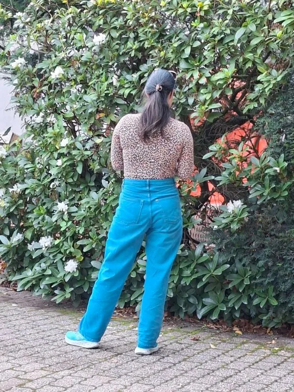 Eine Person mit langen, dunklen Haaren und einer blaue Hose und Oberteil mit Musterdruck. Sie steht auf einem gepflasterten Weg und blickt zu einer dichten, blühenden Hecke. Die Person ist von hinten zu sehen und hält die Arme leicht angewinkelt vor dem Körper.