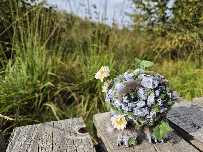 Im Vordergrund steht eine kleine, kugelförmige Figur aus grauen Steinen, Moos und künstlichen Blättern, die mit weißen Blüten dekoriert ist. Die Figur hat zwei grüne Augen und sitzt auf einer hölzernen Oberfläche. Im Hintergrund ist eine dichte, grüne Wiese mit hohen Gräsern und vereinzelten Wildblumen zu sehen. Der Himmel ist hell und leicht bewölkt.