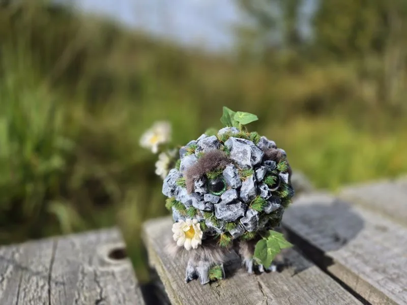 Im Vordergrund steht eine kleine, kugelförmige Figur aus grauen Steinen, Moos und künstlichen Blättern, die mit weißen Blüten dekoriert ist. Die Figur hat zwei grüne Augen und sitzt auf einer hölzernen Oberfläche. Im Hintergrund ist eine unscharfe grüne Landschaft zu sehen. Der Himmel ist hell und leicht bewölkt.