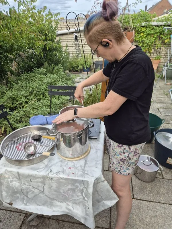 Eine Person mit kurzem, teils gefärbtem Haar steht auf einer Terrasse und rührt mit einem großen Löffel in einem silbernen Topf. Die Person trägt ein schwarzes T-Shirt, gemusterte Shorts und ein Headset. Auf dem Tisch liegen ein großes Metallsieb, ein Kochlöffel und ein blaues Tuch. Im Hintergrund sind Pflanzen, Gartenmöbel und ein gemauerter Zaun zu sehen.