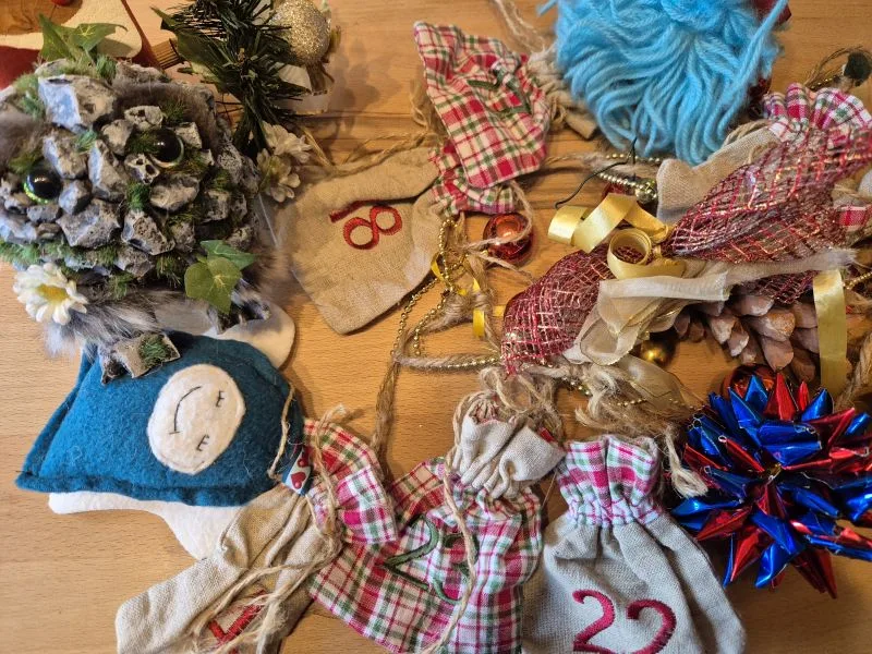 Adventskalender mit kleinen Stoffbeuteln, nummeriert mit roten Ziffern, umgeben von Weihnachtsdekorationen wie Tannenzapfen, Schleifen und einem kleinen blauen Kissen mit Gesicht