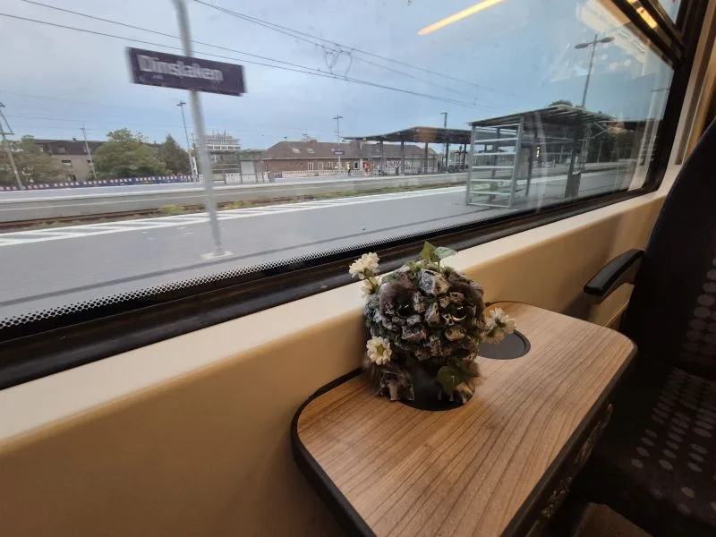 Blick aus einem Zugfenster auf einen Bahnsteig mit Schild 'Dinslaken'. Auf dem Holztisch am Zugfenster sitzt eine kleine, kunstvoll gestaltete Figur. Der Körper besteht aus Steinen, Moos und Pflanzenmaterial. Das Wesen hat große, auffällige Augen. Um den Kopf herum sind kleine weiße Blumen angebracht.