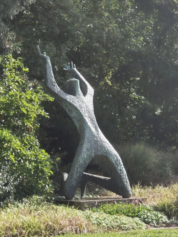 Skulptur einer aufrecht stehenden, abstrakten menschlichen Figur mit erhobenen Armen in einer grünen Parklandschaft