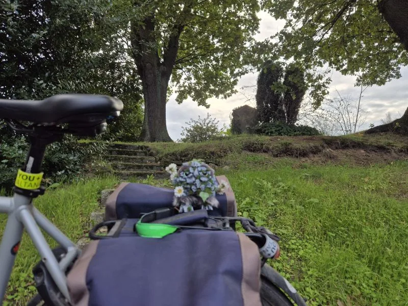Ein Teil eines Fahrrades, Sattel und Gepäcktasche sind vor einer Treppe zu sehen. Die Treppe führt auf einee Wiese mit alten Bäumen. Auf dem Fahrradgepäckträger im Vordergrund sitzt eine kleine, kunstvoll gestaltete Figur. Der Körper besteht aus Steinen, Moos und Pflanzenmaterial. Das Wesen hat große, auffällige Augen. Um den Kopf herum sind kleine weiße Blumen angebracht.