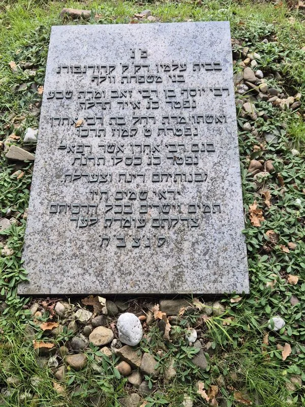 Grabstein mit hebräischer Inschrift auf einem mit Gras und kleinen Steinen bedeckten Boden