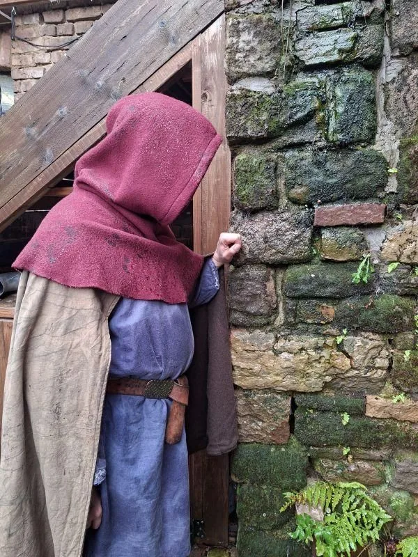 Auf dem Bild sieht man eine Person in mittelalterlich wirkender Kleidung. Die Person trägt eine rote Gugle, und ein langes, blaues Gewand mit einem braunen Gürtel. Darüber liegt ein beiger Umhang. Die Person steht an einer alten Steinwand, die teilweise mit Moos bewachsen ist. Links sieht man eine Holzkonstruktion aus dunklem Holz.